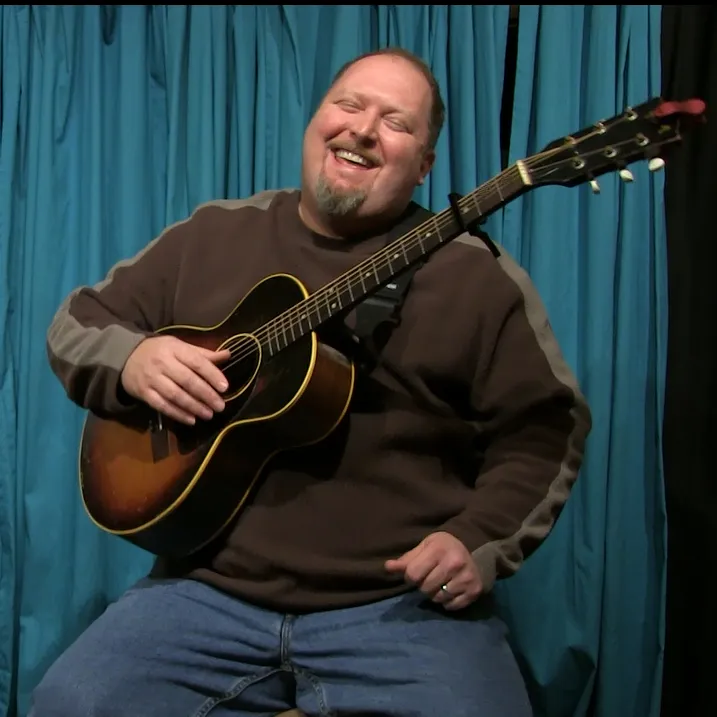 Odie Blackmon laughing mid-song, playing an acoustic guitar in front of a teal stage curtain.